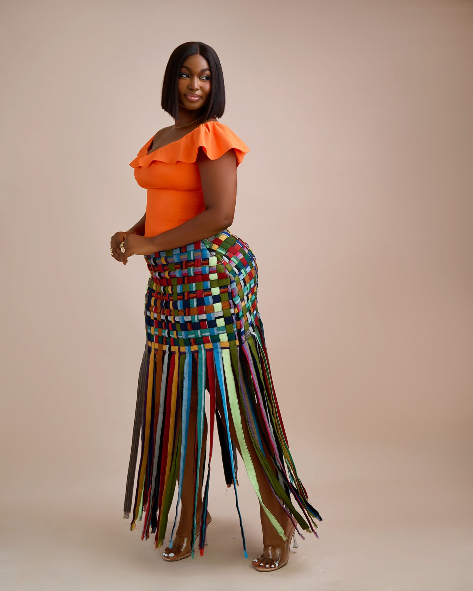Woman wearing an orange top and colorful woven skirt against a beige background