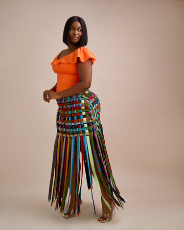 Woman wearing an orange top and colorful woven skirt against a beige background